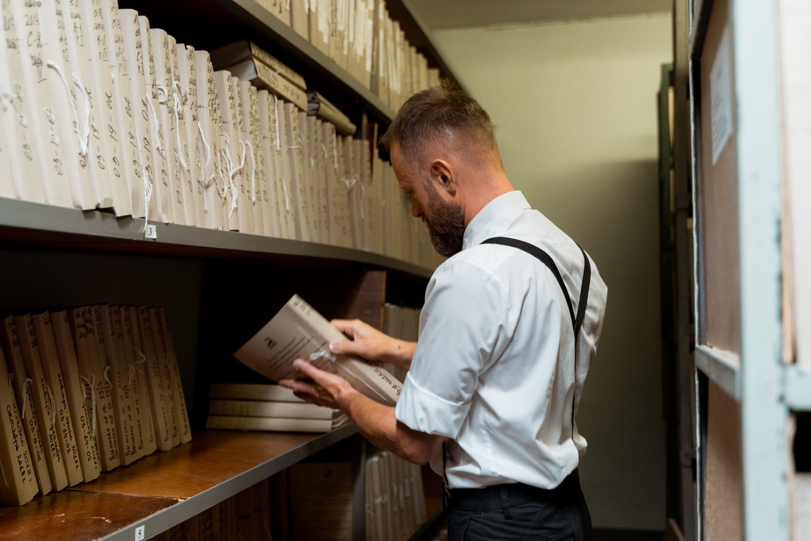 Un homme vêtu de manière décontractée chic organise des dossiers sur des étagères dans une archive.